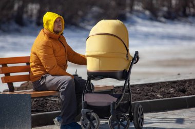 Kapüşonlu bir adam bebek arabasıyla ahşap bankta oturuyor ve...