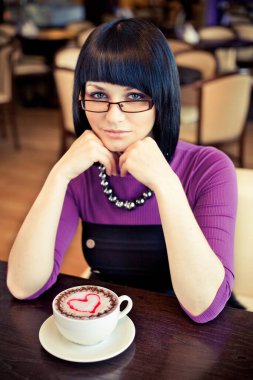 Kafede bir fincan kahveyle oturan gözlüklü çekici bir kadın.