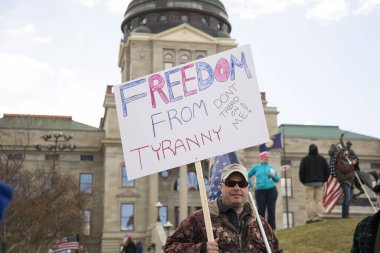 Helena, Montana - April 19, 2020: A man protesting holding a freedom from tyranny sign at a protest at the Capitol of the shutdown of businesses. Capitol building in the background. clipart