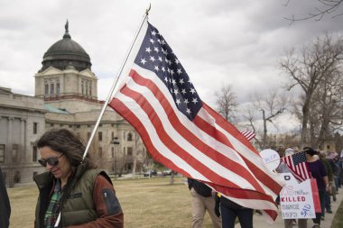 Helena, Montana - 19 Nisan 2020: Elinde bir Amerikan bayrağı olan bir kadın, arkasında hükümet binasının kapatılmasını protesto eden bir grup insanla yürüyor..