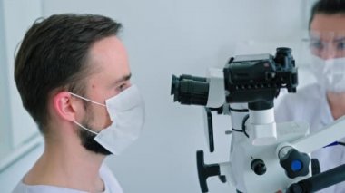 Yüz maskeli diş hekimi diş mikroskobuyla bakıyor.
