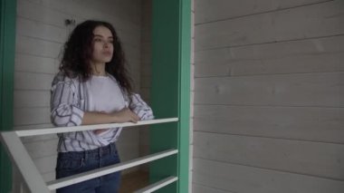 Kotlu ve gömlekli kıvırcık saçlı bir kız güzel bir evin merdivenlerinin yanında duruyor..