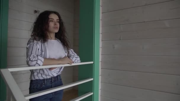 Kręcone włosy dziewczyna w dżinsach i koszuli stoi w pobliżu schodów w pięknym domu. — Wideo stockowe