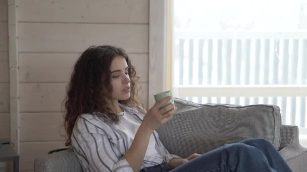 Una chica de pelo rizado en jeans y una camisa bebe café de una taza de abedul en una hermosa casa blanca contra una gran ventana . — Vídeos de Stock