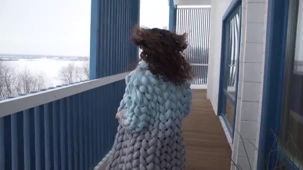 En vacker flicka med lockigt hår i en stor stickad sjal springer runt ett stort hus på vintern och skrattar. — Stockvideo