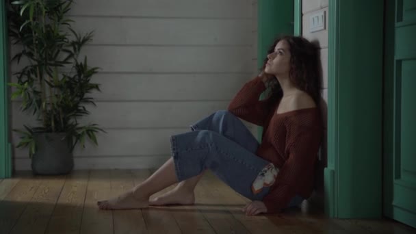 En vacker ung flicka med lockigt hår i jeans och en brun kofta sitter på golvet ser eftertänksamt på vrekh skrattar sedan. — Stockvideo