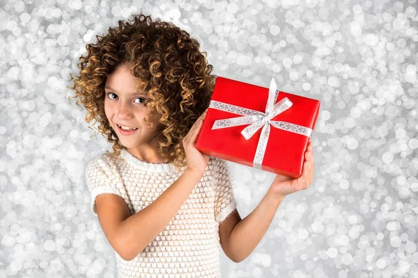 Red Gift. Picture of a little white caucasian girl with curly hair holding red gift box with white ribbon against bright white silver lights background