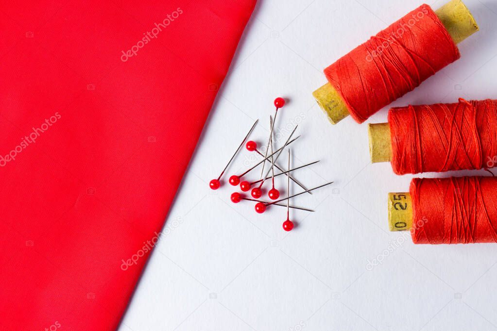 Three spools of scarlet thread. Nearby are needles with scarlet heads. Also nearby is scarlet fabric. Photo on a white background