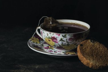 Siyah arka planda çay ve çay poşeti olan bir fincan var. Yakınlarda yuvarlak bir kraker yatıyor.