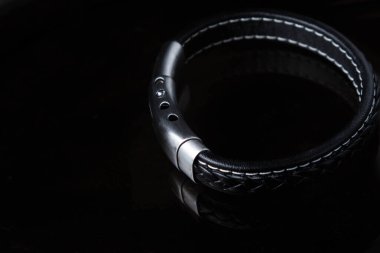 Siyah deri bir erkek bileziği siyah camların üzerinde duruyor. Metal bir kopçası var.