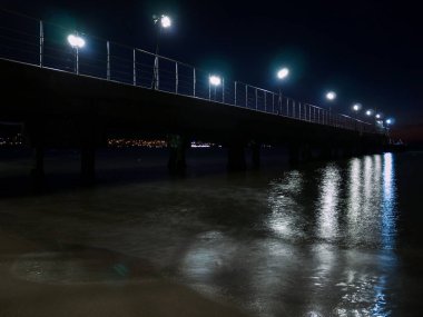 Gece deniz manzarası. İskele fenerlerle aydınlatılıyor. Plaj şemsiyesi ve güneşlik siluetleri..