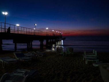 Gece deniz manzarası. İskele fenerlerle aydınlatılıyor. Plaj şemsiyesi ve güneşlik siluetleri..