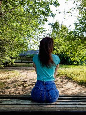 Esmer kız, sırtı kameraya dönük bir şekilde parkta bankta oturuyor. Yeşil tişörtlü ve mavi kot şortlu kız..