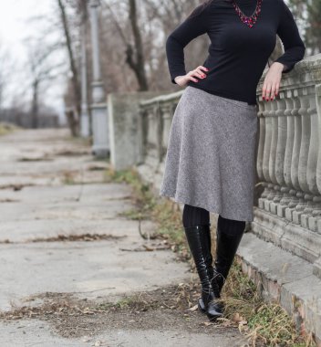 Gri etekli, siyah balıkçı yaka ve siyah deri çizmeli ince bir kadın eski bir sokakta duruyor. kırmızı boncuklar ve oje..