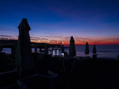 Gece deniz manzarası. İskele fenerlerle aydınlatılıyor. Plaj şemsiyesi ve güneşlik siluetleri..