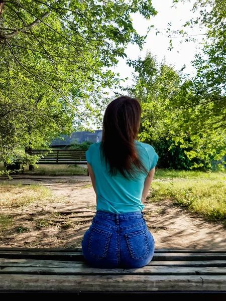 Esmer kız, sırtı kameraya dönük bir şekilde parkta bankta oturuyor. Yeşil tişörtlü ve mavi kot şortlu kız..