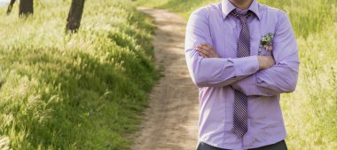 Mor gömlekli ve kravatlı damat ormanda bir patikada duruyor. Ellerini göğsüne bağladı. İş adamı. kendinden emin poz.