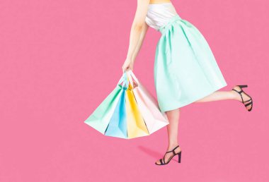 Kadın vücudunun alt tarafı mavi etek ve siyah topuklu ayakkabı giyiyordu. Pembe arka planda birçok pastel renkte alışveriş torbası taşımak