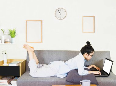 Genç, güzel Asyalı bir kadın mavi gömlek giyer, gri koltukta oturur, online yaşam tarzında çalışır, kahve fincanı ile evde dinlenir, evde çalışır, bilgisayarla çalışır, evden çalışır.