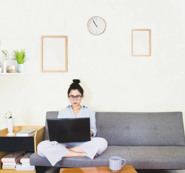 Genç, güzel Asyalı bir kadın mavi gömlek giyer, gri koltukta oturur, online yaşam tarzında çalışır, kahve fincanı ile evde dinlenir, evde çalışır, bilgisayarla çalışır, evden çalışır.