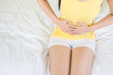 Sarı atlet giyen genç Asyalı kadın pencerenin yanındaki beyaz yatakta oturuyor. İnce perdeli, karın ağrısı çeken bir kadının el ele tutuşması. Karın ağrısı konsepti, seçici bir odak noktası.