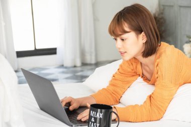 Genç, güzel Asyalı bir kadın portakal rengi örgü giyer, yatağa uzanır, dizüstü bilgisayarla oynar. Ev bilgisayarı, online yaşam tarzı, oda rahatlığı, iş hayatı dengesi, ev kahvesi kupası, ev işi, ev işi.