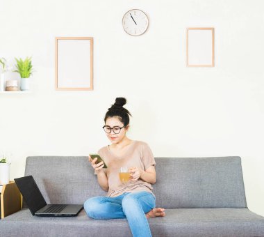 Genç, güzel Asyalı bir kadın kahverengi tişört giyer ve kot pantolon giyer, gri koltukta yatar ve oturur, online yaşam tarzında çalışır, kahve fincanı ile evde dinlenir, evde çalışır, bilgisayarla çalışır, evden çalışırdı.