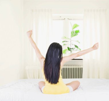  Sarı fanila giyen genç Asyalı kadın yoga yaparken ince perdeli beyaz bir yatakta otururken, evde sağlık hizmetleri konseptinde uyandıktan sonra uzanıyor.