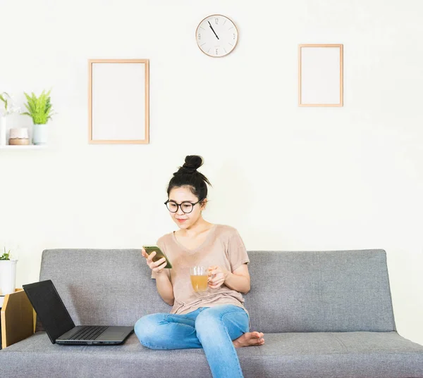 Genç, güzel Asyalı bir kadın kahverengi tişört giyer ve kot pantolon giyer, gri koltukta yatar ve oturur, online yaşam tarzında çalışır, kahve fincanı ile evde dinlenir, evde çalışır, bilgisayarla çalışır, evden çalışırdı.