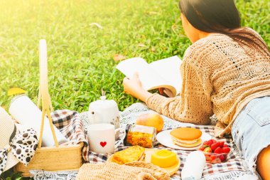 Piknik konsepti, genç, güzel Asyalı kadın çimenlerde oturup kitap okuyor, tatillerde eğleniyor, seçmeli odaklanıyor.