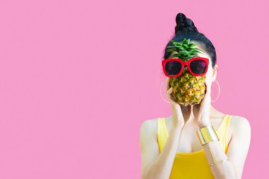 Sarı fanilalı mutlu Asyalı kadın, güneş gözlüğü modası elinde ananas, yaz tatillerinde ananaslı komik kadın pembe arka planda izole.