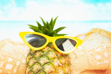 Plaj, kırmızı güneş gözlüklü komik ananas ve deniz ve gökyüzüne karşı kumun üzerinde şapka. Tropik yaz tatili konsepti. Kumsalda güneşli bir gün seçici odak noktası sarı ve mavi deniz.