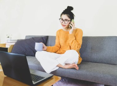 yumuşak seçici odak, güzel Asyalı kadın turuncu örgü giyer, gri koltukta oturur, online yaşam tarzında çalışır, evde kahve içer, ev bilgisayarında çalışır, evden çalışır.