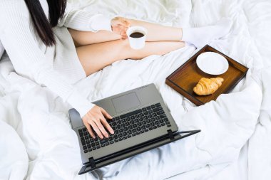 Genç Asyalı kadınların yatakta beyaz örgü elbiseler giyip, yatak odasından dizüstü bilgisayarla çalışıp, internet üzerinden çalışıp, evde kahve içip, seçici bir odak noktasıyla rahatlayışının en üst görüntüsü.