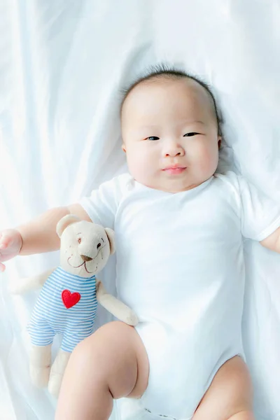 top view Portrait of a newborn Asian cute baby boy wore white Infant bodysuit on the bed , Charming Fat baby 5 month old lies in bed with a doll,enjoy and happy,soft selective focus