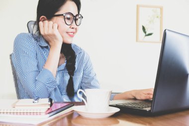 Seçici odak noktası, gülümseyen güzel Asyalı kadın gözlük takıyordu, mavi çizgili gömlek masada oturuyordu, online yaşam tarzı üzerinde çalışıyordu kahveyle evde dinleniyor, dizüstü bilgisayarla çalışıyor, ev konseptinden çalışıyor.