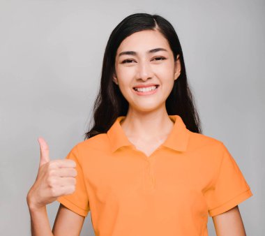 Genç ve güzel Asyalı bir kadın, uzun siyah saçlı, turuncu tişört giyiyor, gülümsüyor ve kendinden emin, harika bir başparmağı var.