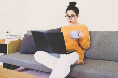 Genç, güzel Asyalı bir kadın turuncu örgü giyer, gri koltukta oturur, online yaşam tarzında çalışır, kahve fincanı ile evde dinlenir, bilgisayarla çalışır, evden çalışır.