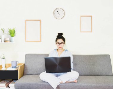 Genç, güzel Asyalı bir kadın mavi gömlek giyer, gri koltukta oturur, online yaşam tarzında çalışır, kahve fincanı ile evde dinlenir, evde çalışır, bilgisayarla çalışır, evden çalışır.