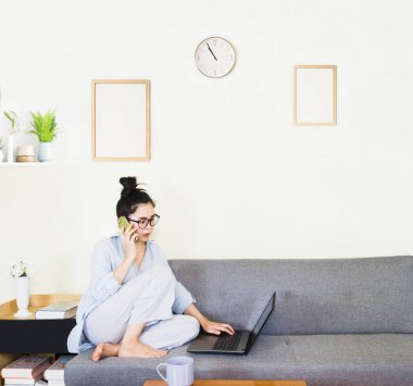 Genç, güzel Asyalı bir kadın mavi gömlek giyer, gri koltukta oturur, online yaşam tarzında çalışır, kahve fincanı ile evde dinlenir, evde çalışır, bilgisayarla çalışır, evden çalışır.