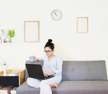 Genç, güzel Asyalı bir kadın mavi gömlek giyer, gri koltukta oturur, online yaşam tarzında çalışır, kahve fincanı ile evde dinlenir, evde çalışır, bilgisayarla çalışır, evden çalışır.