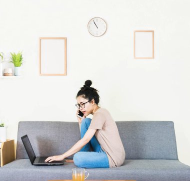 Genç, güzel Asyalı bir kadın kahverengi tişört giyer ve kot pantolon giyer, gri koltukta yatar ve oturur, online yaşam tarzında çalışır, kahve fincanı ile evde dinlenir, evde çalışır, bilgisayarla çalışır, evden çalışırdı.