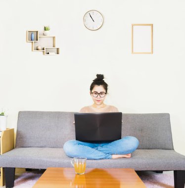 Genç, güzel Asyalı bir kadın kahverengi tişört giyer ve kot pantolon giyer, gri koltukta yatar ve oturur, online yaşam tarzında çalışır, kahve fincanı ile evde dinlenir, evde çalışır, bilgisayarla çalışır, evden çalışırdı.
