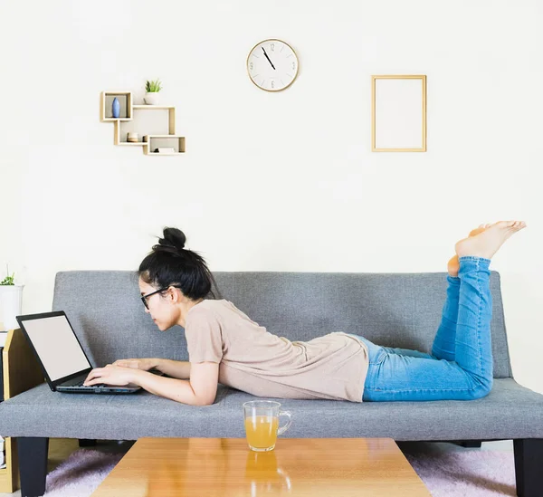 Genç, güzel Asyalı bir kadın kahverengi tişört giyer ve kot pantolon giyer, gri koltukta yatar ve oturur, online yaşam tarzında çalışır, kahve fincanı ile evde dinlenir, evde çalışır, bilgisayarla çalışır, evden çalışırdı.