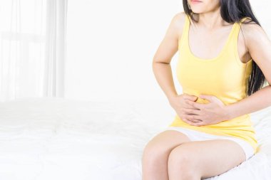 Sarı fanila giyen genç Asyalı kadın pencerenin yanındaki beyaz yatakta oturuyor. İnce perdeli, karın ağrısı, karın ağrısı, sağlık sigortası konsepti olan kadın el ele tutuşuyor.