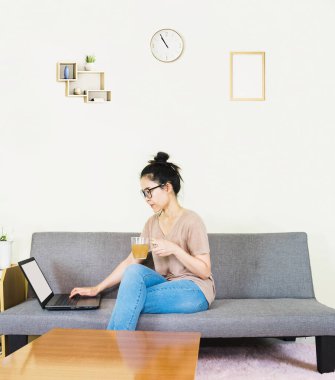 Genç, güzel Asyalı bir kadın kahverengi tişört giyer ve kot pantolon giyer, gri koltukta yatar ve oturur, online yaşam tarzında çalışır, kahve fincanı ile evde dinlenir, evde çalışır, bilgisayarla çalışır, evden çalışırdı.