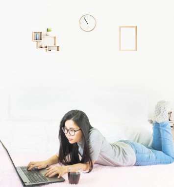 Genç, güzel Asyalı bir kadın gri tişört giyer ve kot pantolon giyer, oturma odasında pembe halıya uzanır, online yaşam tarzında çalışır, kahve fincanıyla evde dinlenir, dizüstü bilgisayarla çalışır, evden çalışırdı.