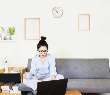 Genç, güzel Asyalı bir kadın mavi gömlek giyer, gri koltukta oturur, online yaşam tarzında çalışır, kahve fincanı ile evde dinlenir, evde çalışır, bilgisayarla çalışır, evden çalışır.