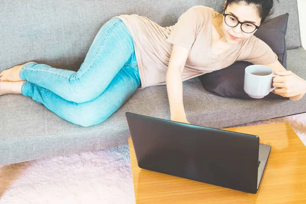 Young beautiful asian woman wore brown t shirt and jean, lying and sitting on gray sofa ,work online lifestyle,relax at home with coffee mug,home working laptop,work from home