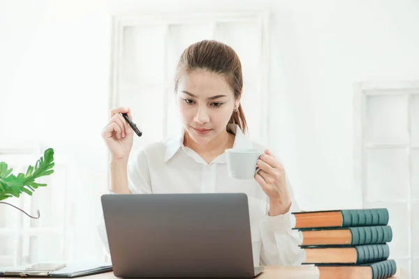 young Beautiful asian business woman wearing white shirt, sitting on work dest. work from home with laptop and relax with coffee